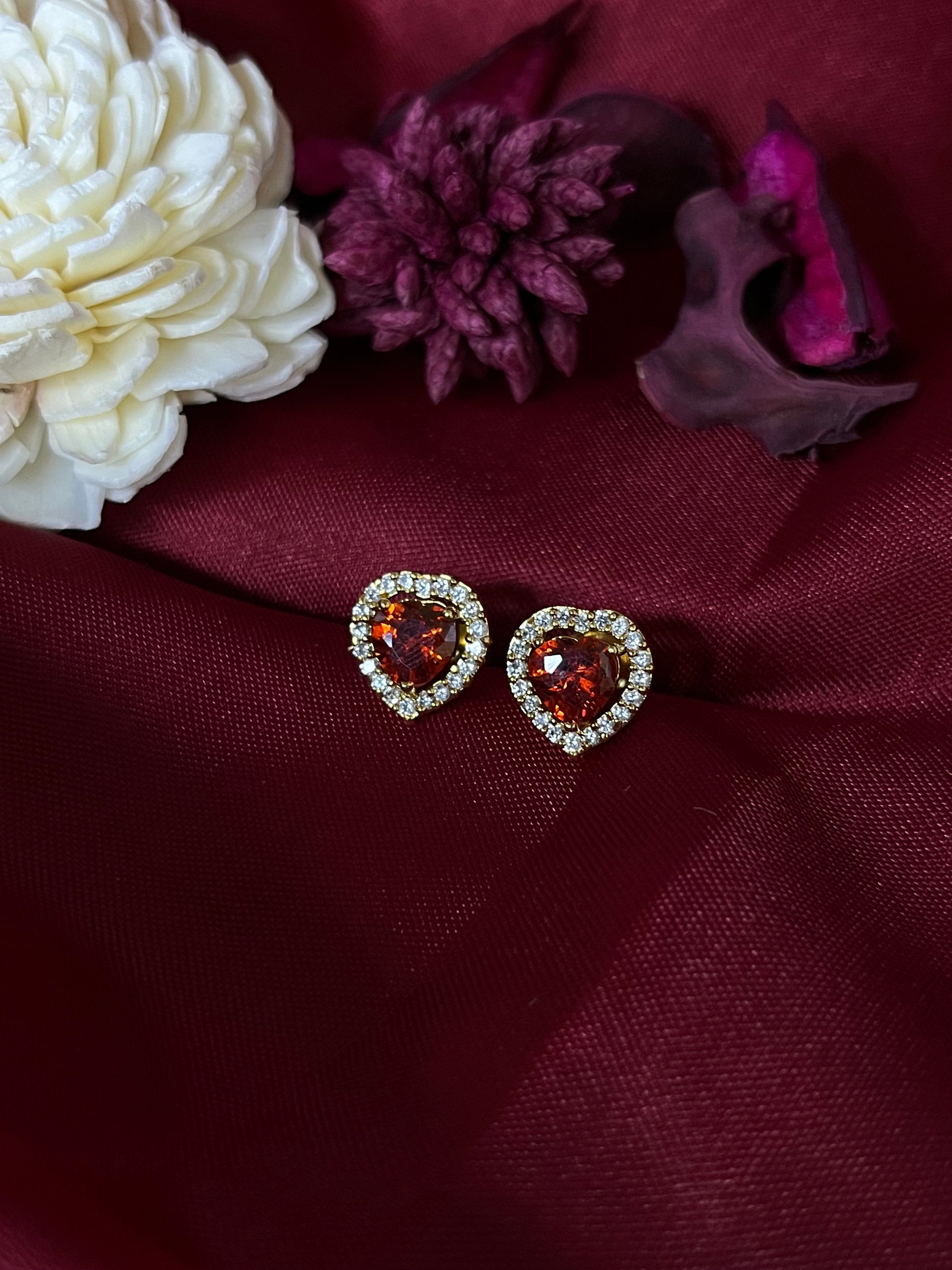 Heart-shaped earrings with red gemstones on a burgundy fabric background with flowers.